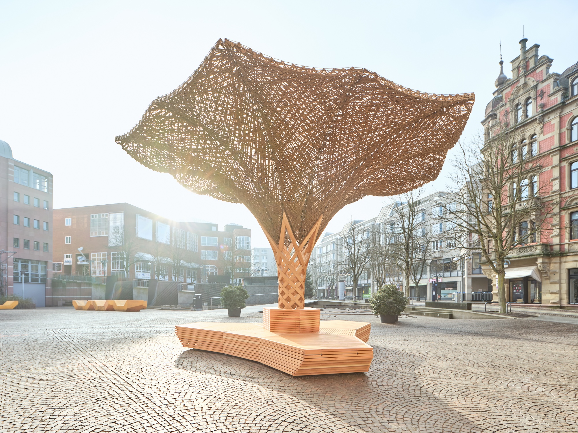 Deutsch: komorebi Installation auf dem Kronenplatz in Karlsruhe, eine organisch geformte Holzstruktur mit geflochtenem Sonnendach aus Naturfasern, die als öffentlicher Sitz- und Aufenthaltsort dient. English: komorebi installation on Kronenplatz in Karlsruhe, featuring an organically shaped timber structure with a woven natural-fiber canopy serving as a public seating and gathering space.
